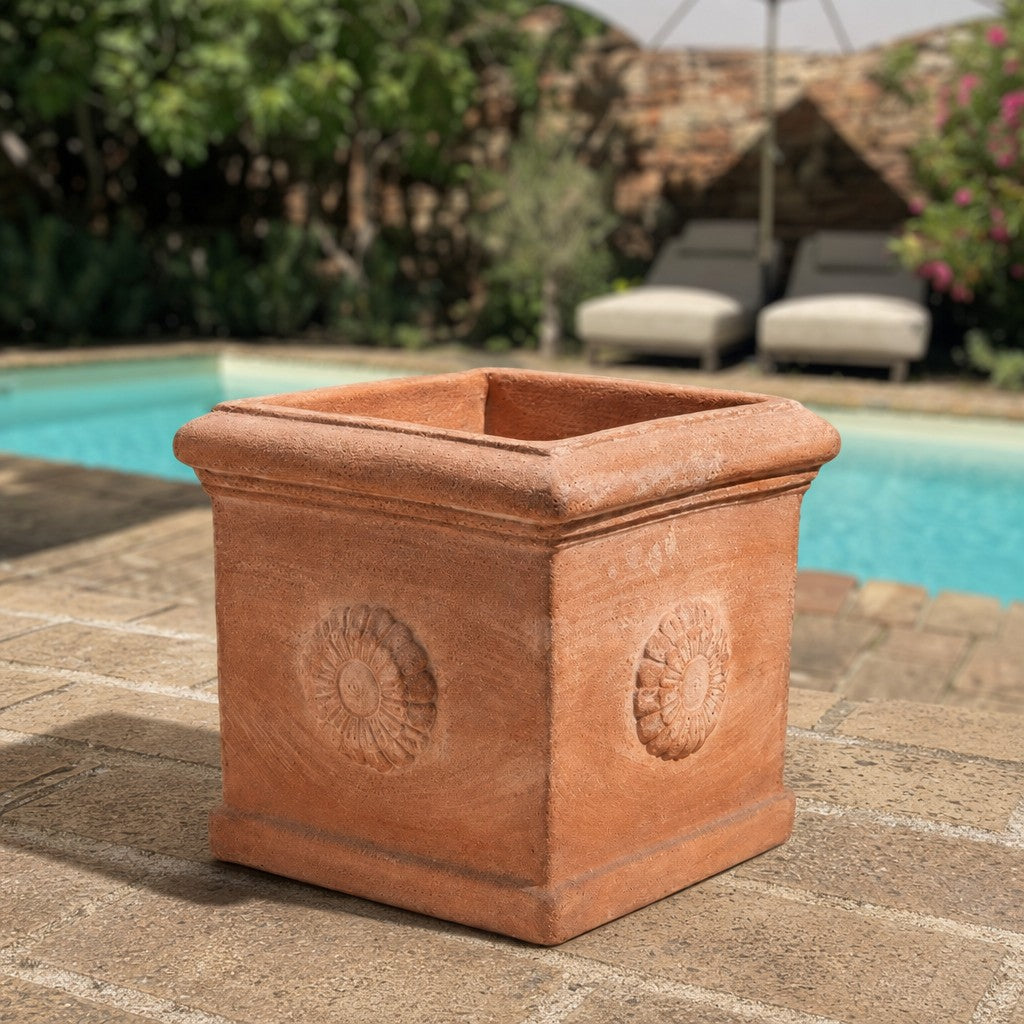 Der quadratische Terrakotta-Kübel „Rosone“ mit seinem floralen Muster steht auf einer gemauerten Terrasse neben dem Swimmingpool, umgeben von Liegestühlen und üppigem Grün im Hintergrund.