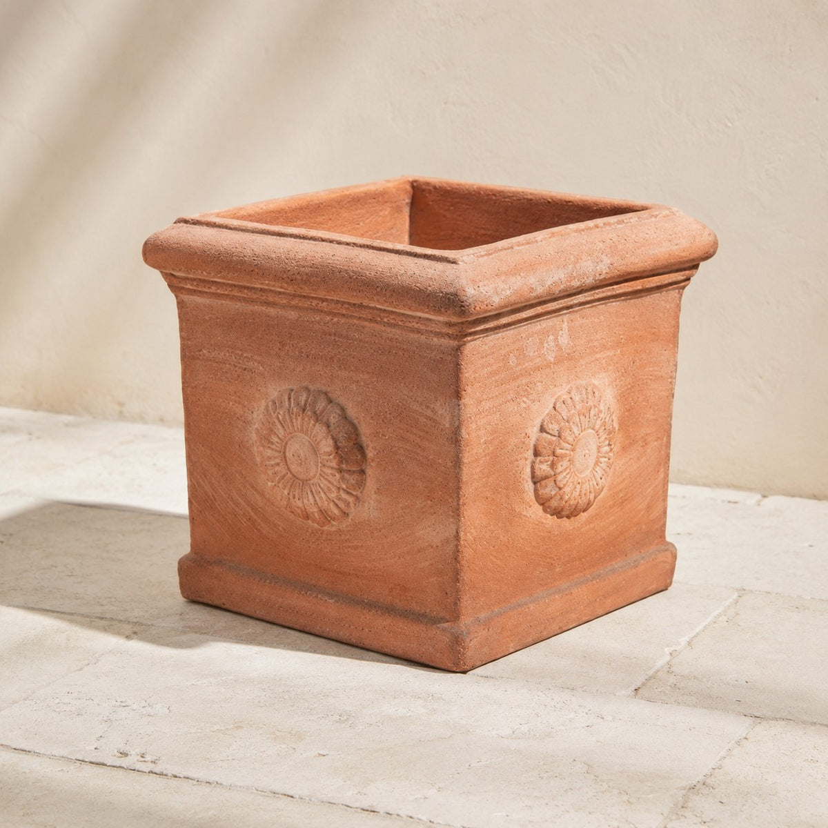 Ein quadratischer Terrakotta-Kübel „Rosone“ mit erhabenen Blumenmedaillons an jeder Seite steht auf einem hellen Steinboden vor einer beigen Wand.