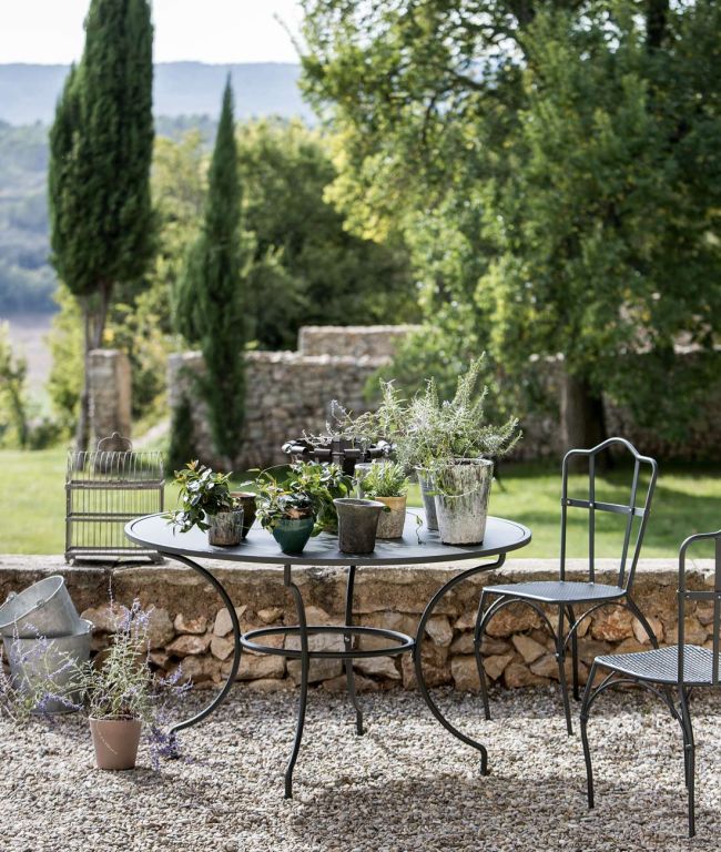 In einer ruhigen Gartenumgebung im Freien steht ein runder Tisch Toscana (125 cm), auf dem verschiedene Topfpflanzen in einem Kieshof stehen. Zwei passende Metallstühle stehen daneben. Im Hintergrund sind üppige Bäume, eine Steinmauer und in der Ferne Hügel unter einem klaren Himmel zu sehen.