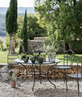 In einer ruhigen Gartenumgebung im Freien steht ein runder Tisch Toscana (125 cm), auf dem verschiedene Topfpflanzen in einem Kieshof stehen. Zwei passende Metallstühle stehen daneben. Im Hintergrund sind üppige Bäume, eine Steinmauer und in der Ferne Hügel unter einem klaren Himmel zu sehen.