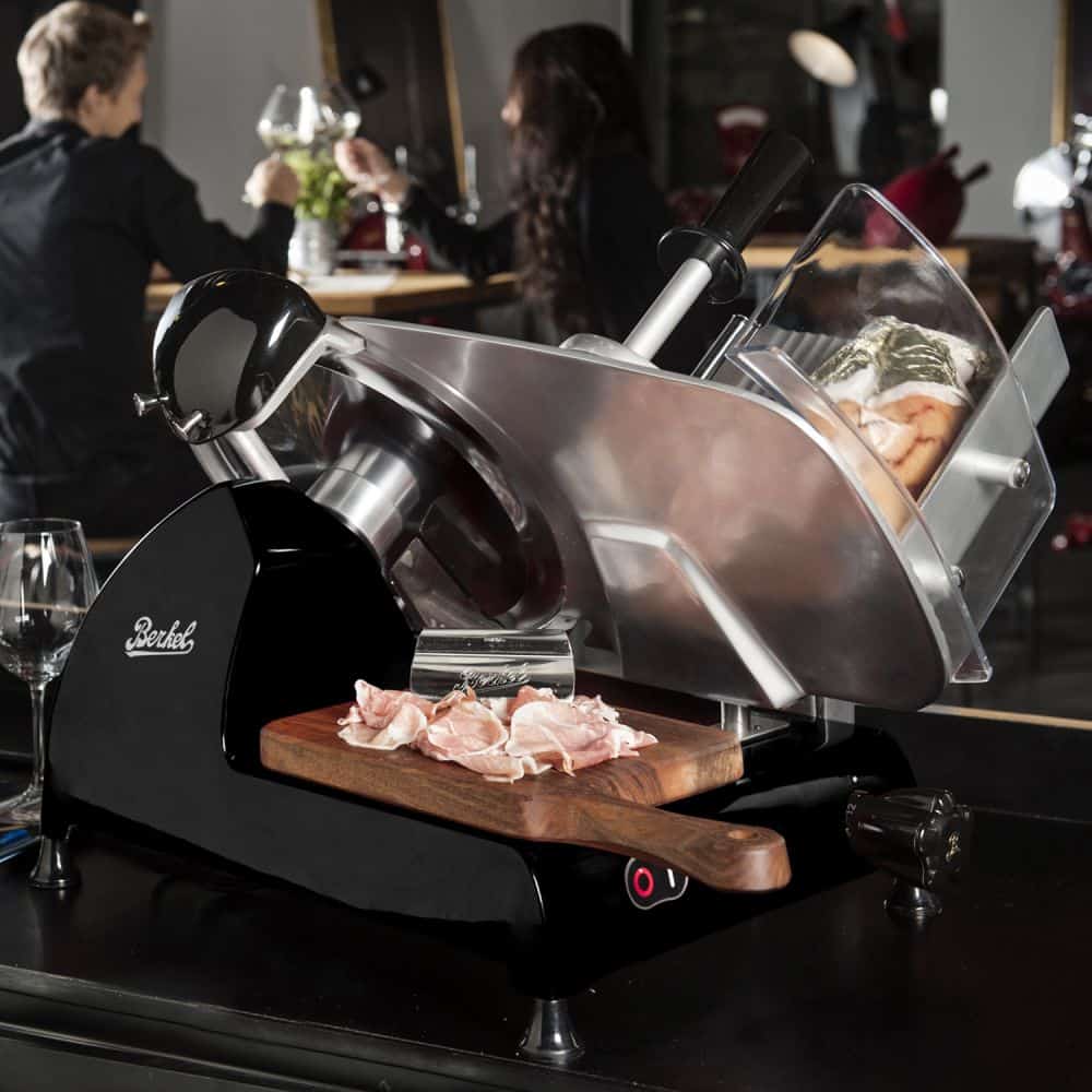 In einer Küchenumgebung wird eine Berkel Red Line 300 Schwarz Original Profi-Aufschnittmaschine gezeigt, die aktiv Fleisch schneidet. Auf dem hölzernen Fassholz-Einlegebrett unter der Aufschnittmaschine sind mehrere dünne Scheiben Schinken zu sehen. Mit seinem beeindruckenden Messerdurchmesser von 300 mm liefert dieses Modell Präzision. Im Hintergrund sind unscharf zwei Personen zu sehen, die miteinander interagieren.