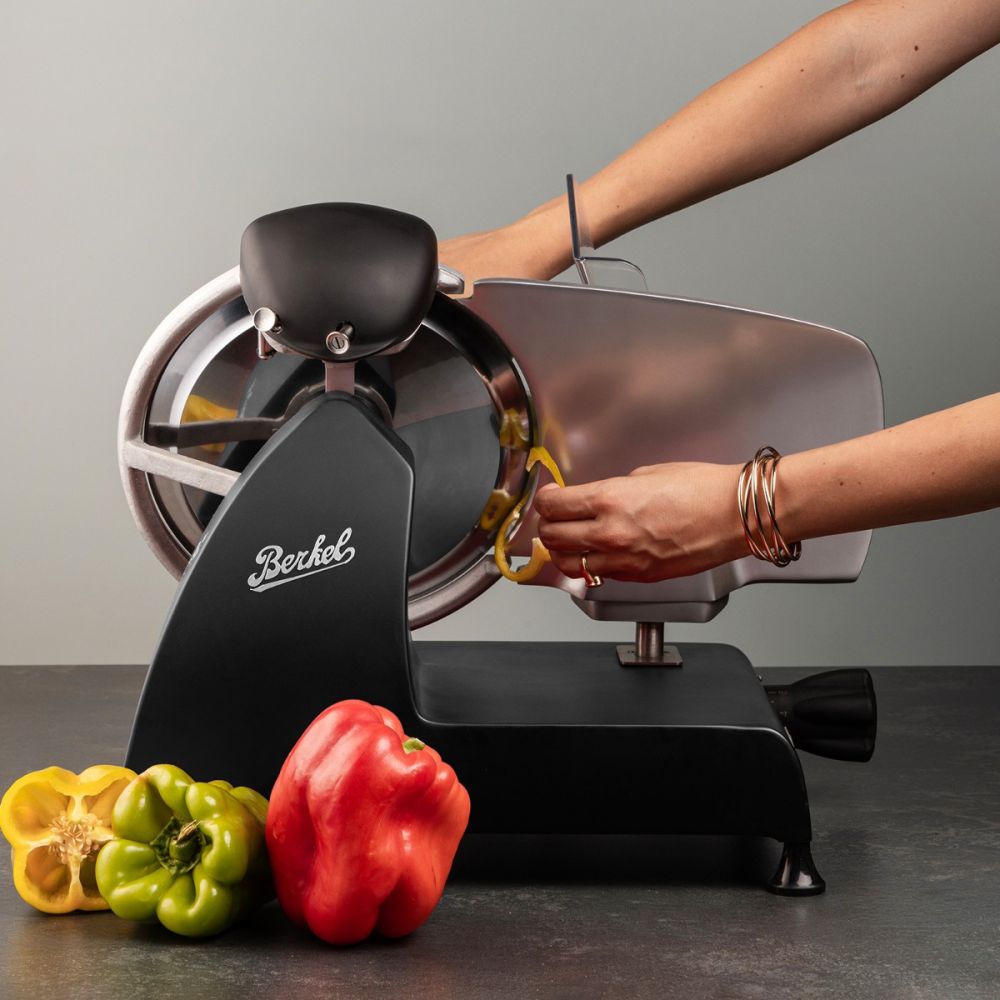 Mit dem Berkel RedLine 250 MATT schwarz mit Fassholzbrett schneidet jemand eine gelbe Paprika, während daneben grüne und rote Paprika auf der dunklen, neutralen Arbeitsplatte liegen.