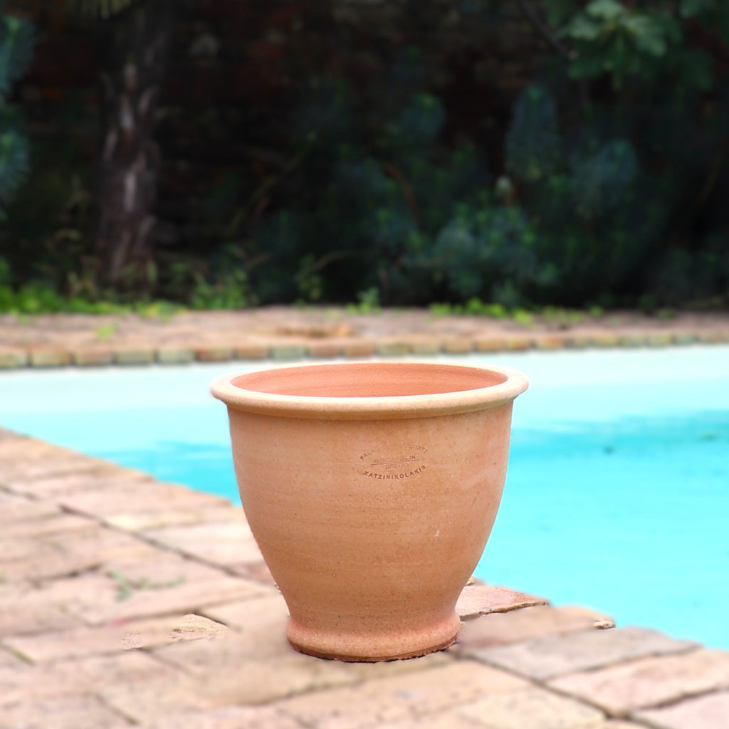 Ein großer, leerer Terrakotta-Pflanztopf "Rubus" 30 cm - Handgemacht & Frostfest steht am Rande einer gemauerten Terrasse an einem blauen Swimmingpool, mit üppigem Grün und einer Backsteinmauer im unscharfen Hintergrund.