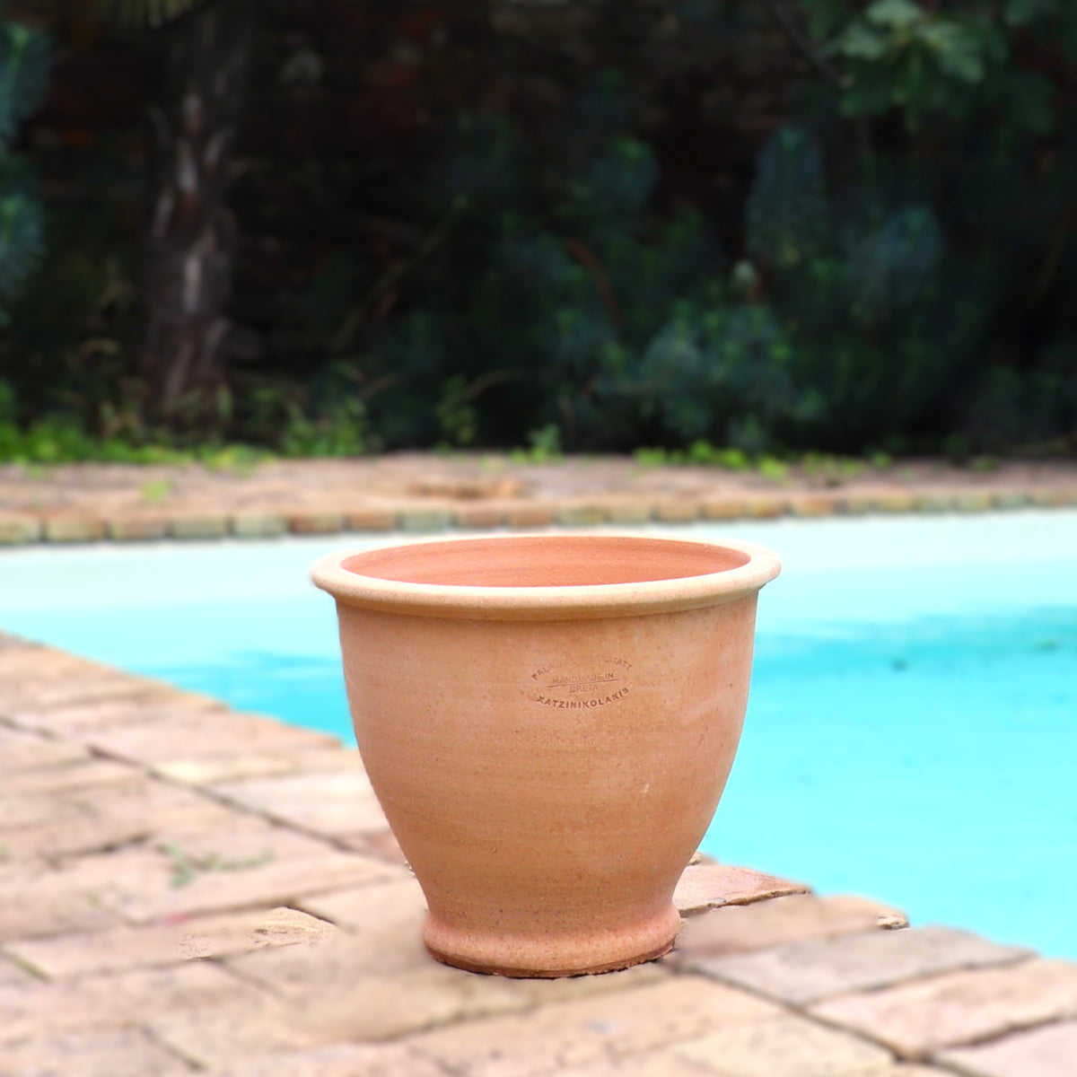 Ein großer, leerer Terrakotta-Pflanztopf "Rubus" 30 cm - Handgemacht & Frostfest steht am Rande einer gemauerten Terrasse an einem blauen Swimmingpool, mit üppigem Grün und einer Backsteinmauer im unscharfen Hintergrund.