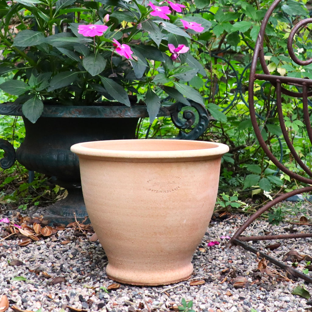 Ein großer, leerer Terrakotta Pflanztopf "Rubus" 30 cm - handgemacht & frostfest - steht auf Kies im Garten. Dahinter befindet sich ein schwarzer Topf mit blühenden pinken Blumen; rechts ist eine Metall-Gartendeko zu sehen.