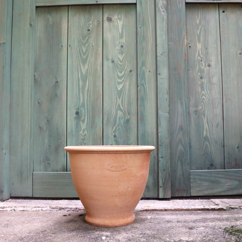 Ein Terrakotta Pflanztopf "Rubus" 30 cm - Handgemacht & Frostfest steht leer auf einer Betonfläche vor einer verwitterten grünen Holztür.