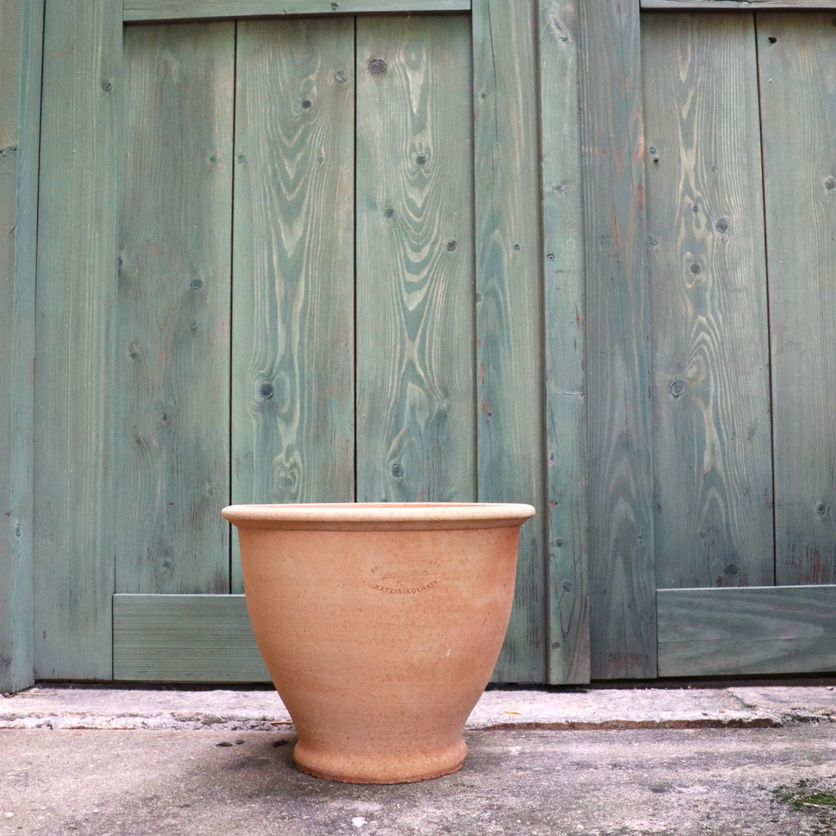 Ein Terrakotta Pflanztopf "Rubus" 30 cm - Handgemacht & Frostfest steht leer auf einer Betonfläche vor einer verwitterten grünen Holztür.