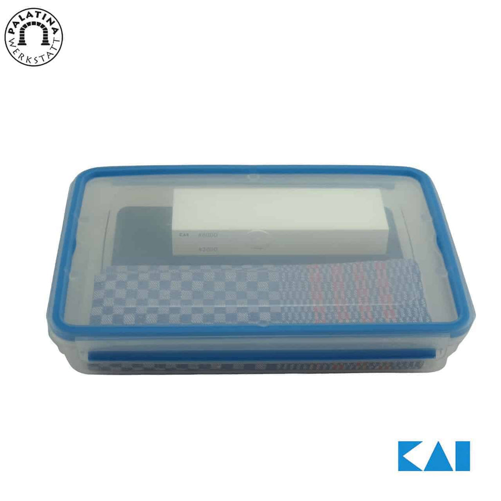 Eine rechteckige, transparente Plastikbox mit blauem Rand, die einen weißen Artikel und zwei karierte Tücher enthält. Die Box zeigt das Kai-Logo in der unteren rechten Ecke und ein rundes Logo mit dem Text „Kalatma Yeksta“ in der oberen linken Ecke. Es scheint Teil des hochwertigen 4-teiligen Schleifstein- und Pflegesets AP-0316 von Kai zu sein.