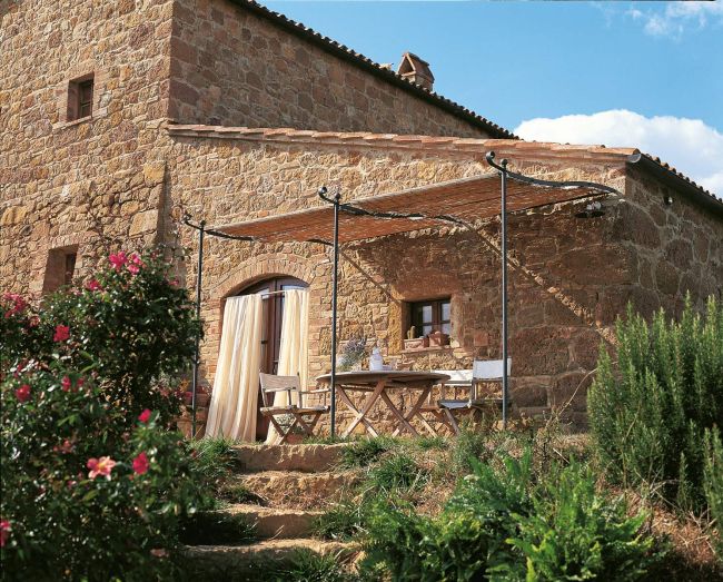 Steinhaus mit einem hölzernen Vordach über einer Terrasse mit Holztisch und Stühlen. Die Terrasse ist von üppigem Grün und blühenden Blumen umgeben. An der Hauswand sind unter einer gewölbten Anbaupergola Solaire ein kleines Fenster und eine gewölbte Tür mit Vorhang zu sehen. Der Himmel ist teilweise bewölkt.