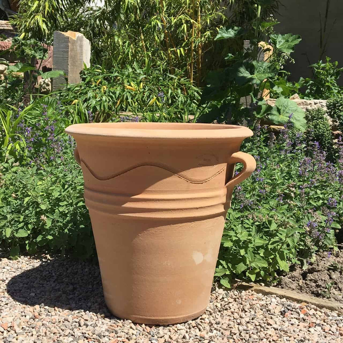 In einem Garten steht ein großer Terrakotta-Pflanztopf, Blumenkübel „Salvia“, mit Griffen auf beiden Seiten auf einem Kiesweg. Dieser 34 cm hohe Pflanzkübel ist von üppigen grünen Pflanzen und Blumen umgeben, im Hintergrund sind Blattwerk und eine Gartenmauer zu sehen. Salvia-Blüten sind zwischen dem kräftigen Grün verstreut.