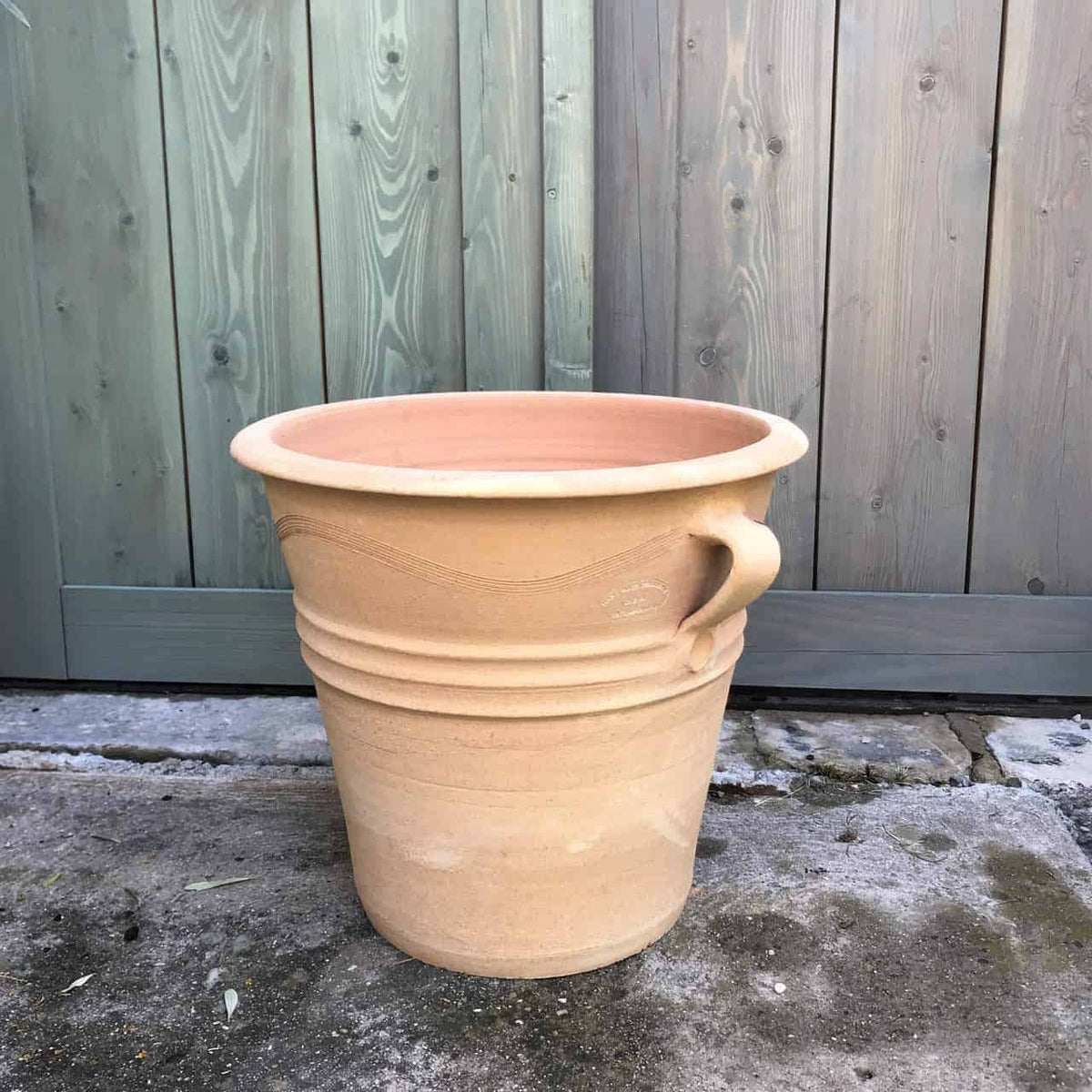 Ein großer, beigefarbener Terrakotta-Pflanztopf, Blumenkübel „Salvia“, mit einer Höhe von 34 cm steht auf einer Betonfläche vor einem Holzzaun. Der Topf hat ein einfaches, unglasiertes Design mit einem einzelnen gebogenen Griff und dezenten dekorativen horizontalen Rillen in der Mitte, perfekt für die Präsentation leuchtender Salvia-Blüten.
