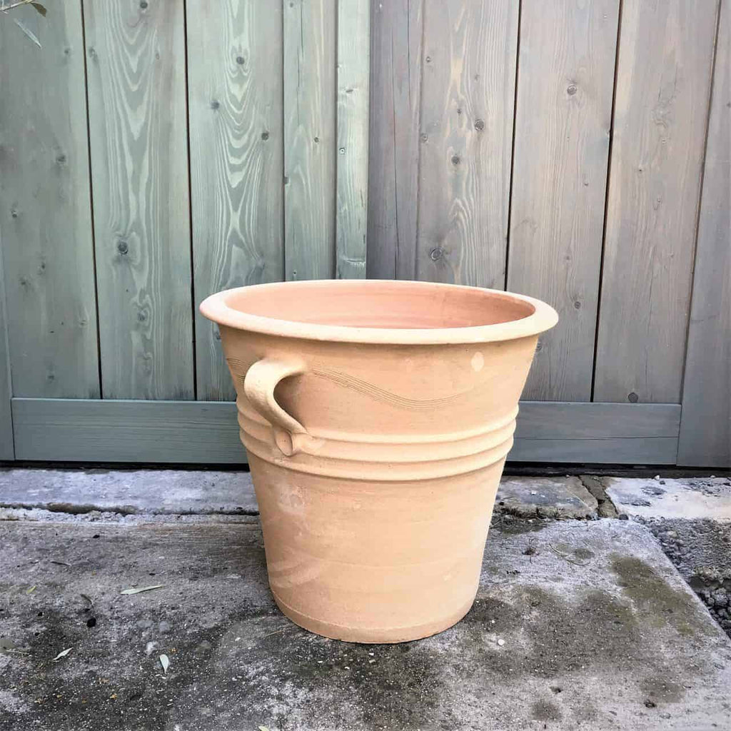 Ein großer, leerer Terrakotta-Pflanztopf Blumenkübel „Salvia“, 34 cm hoch, mit dekorativem Griff und steht auf Betonboden vor einem verwitterten Holzzaunhintergrund. Der Topf weist ein einfaches, rustikales Design mit zwei konzentrischen Rillen um die breiteste Stelle auf und ist damit perfekt zum Pflanzen von leuchtendem Salbei oder anderen Blumen in Ihrem Garten.