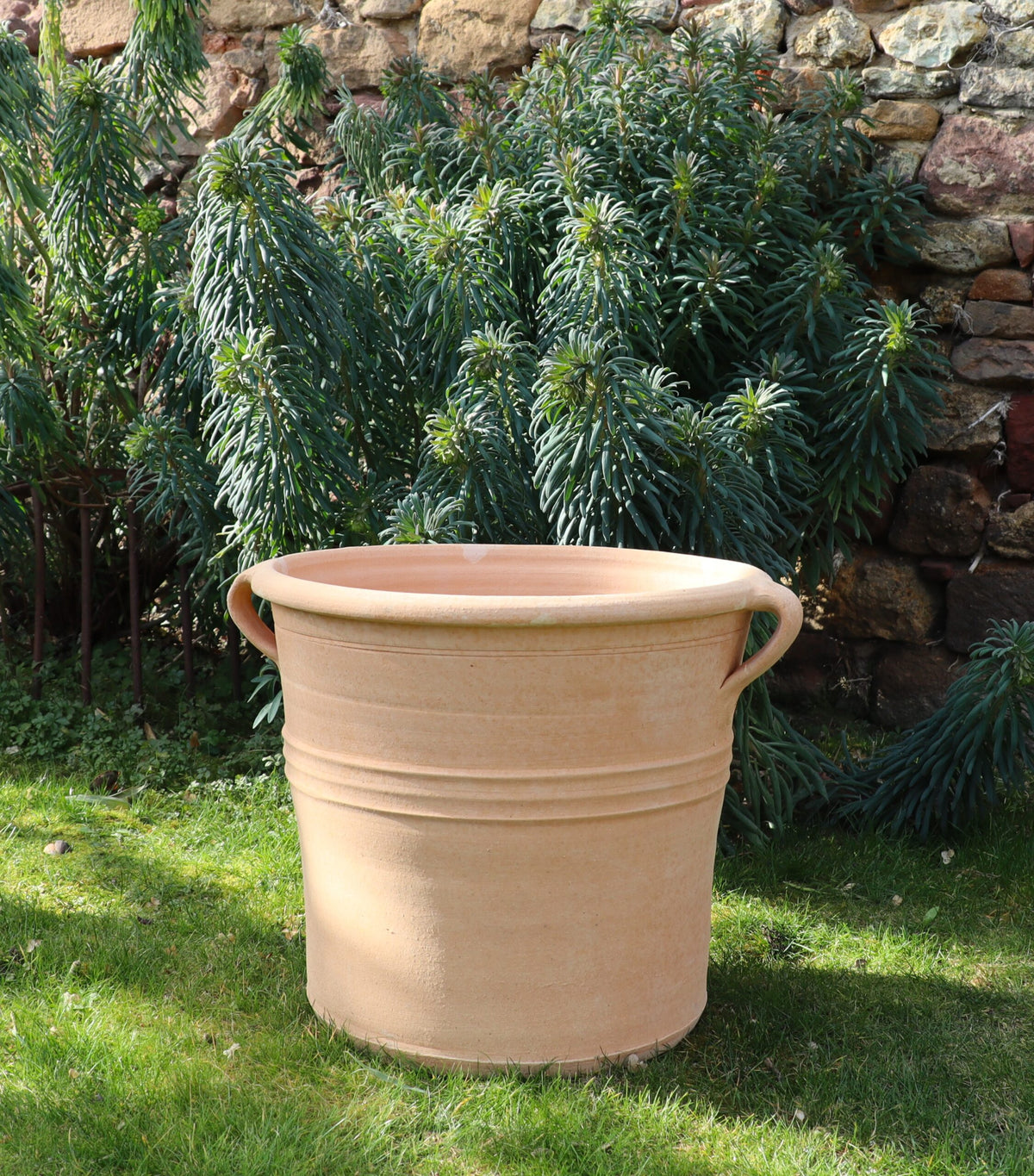 Der Pflanzkübel „Salvia 2“, H=40 cm, ein großer Terrakotta-Pflanzkübel mit Griffen an beiden Seiten, steht leer auf einer Rasenfläche vor einem üppigen grünen Busch und einer rustikalen Steinmauer. Mit seinem schlichten Streifendesign wartet der gut beleuchtete Pflanzkübel darauf, gefüllt zu werden, vielleicht mit leuchtendem Salvia.