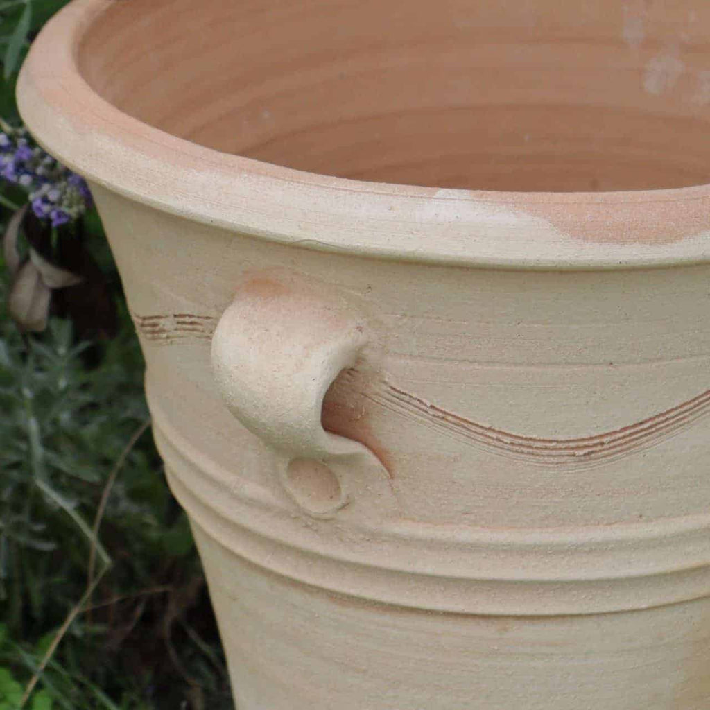 Dieses Bild zeigt eine Nahaufnahme des Terrakotta-Pflanztopfs, Blumenkübel „Salvia“, H=34 cm, und zeigt seine Größe und seinen beigen Farbton. Der Topf hat einen einzelnen Griff und eine glatte, aber leicht geriffelte Oberfläche mit minimaler Verzierung. Der Hintergrund enthält einige grüne Blätter und violette Blumen, was an eine Gartenumgebung im Freien erinnert.