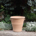 Ein großer, beigefarbener, winterfester Blumentopf „Salvia“, H=40 cm, aus Terrakotta mit zwei Griffen auf jeder Seite steht auf einer Betonfläche, umgeben von weiß blühenden Pflanzen und grünem Blattwerk. Der Topf weist ein schlichtes Design mit horizontalen Linien und einem Wellenmuster am oberen Rand auf, ergänzt durch Salvia-Blüten in der Nähe.