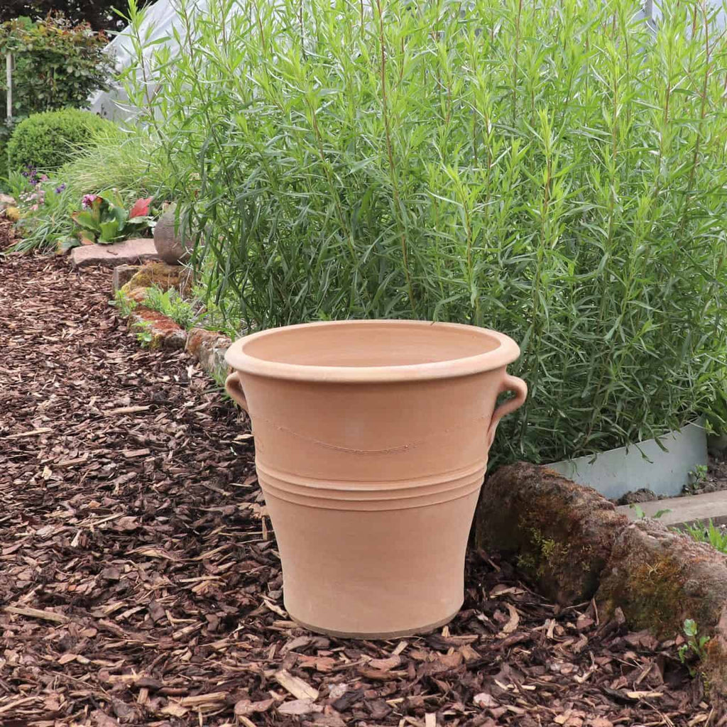 Ein winterfester Blumentopf „Salvia“, 40 cm hoch und mit zwei seitlichen Henkeln versehen, steht auf einem braunen Mulchweg in einem Garten. Hinter dem Topf wachsen hohe Grünpflanzen und üppiger Salvia, der Weg ist mit Steinen und anderen Pflanzen gesäumt. Der Garten wirkt üppig und gepflegt, wodurch der Topf schön zur Geltung kommt.