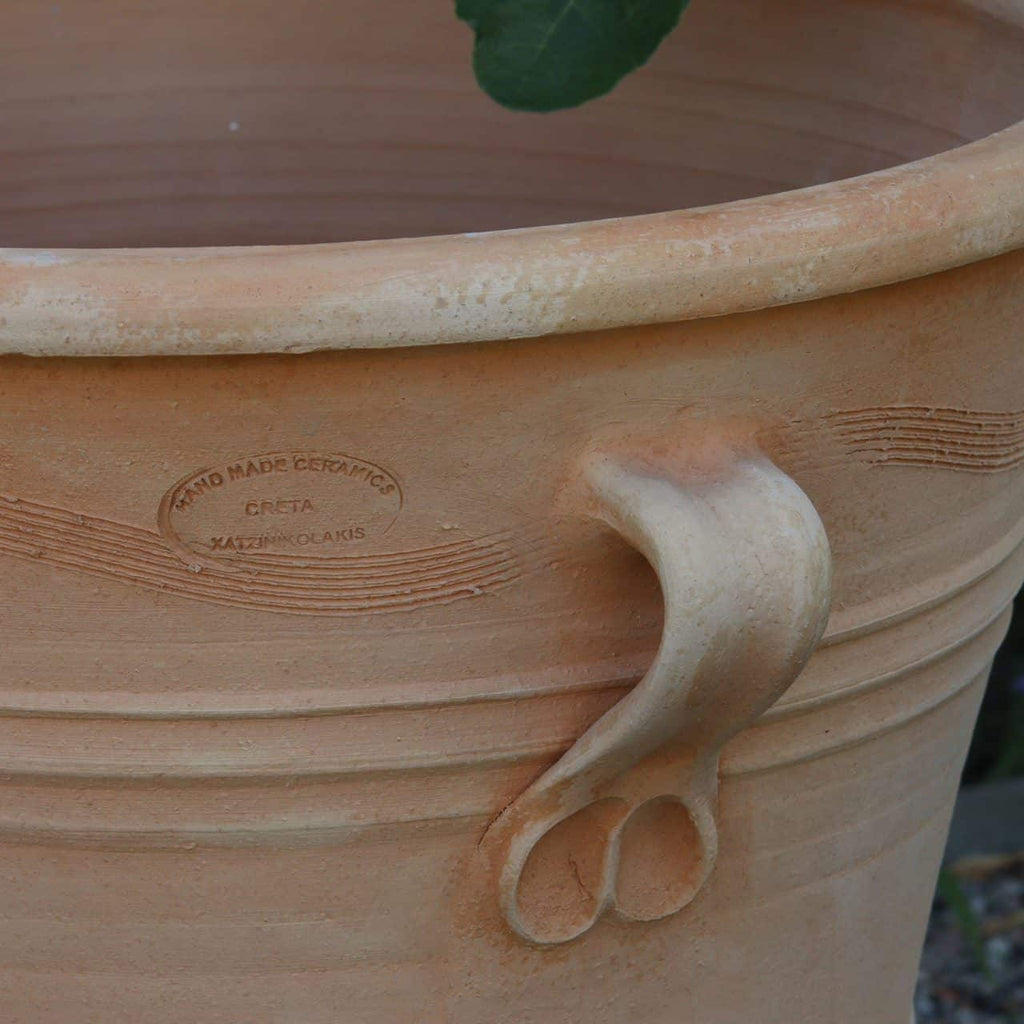 Eine Nahaufnahme eines großen, frostfesten Blumenkübels aus Terrakotta „Salvia“, 50 cm hoch, mit strukturiertem Griff. Der Topf trägt an der Seite das Logo mit der Aufschrift „HAND MADE CERAMIC CRETA JAMSINIOULAKIS“. Oben ist ein Teil einer grünen Salvia-Pflanze zu sehen, die in ihrem Pflanzgefäß gedeiht.