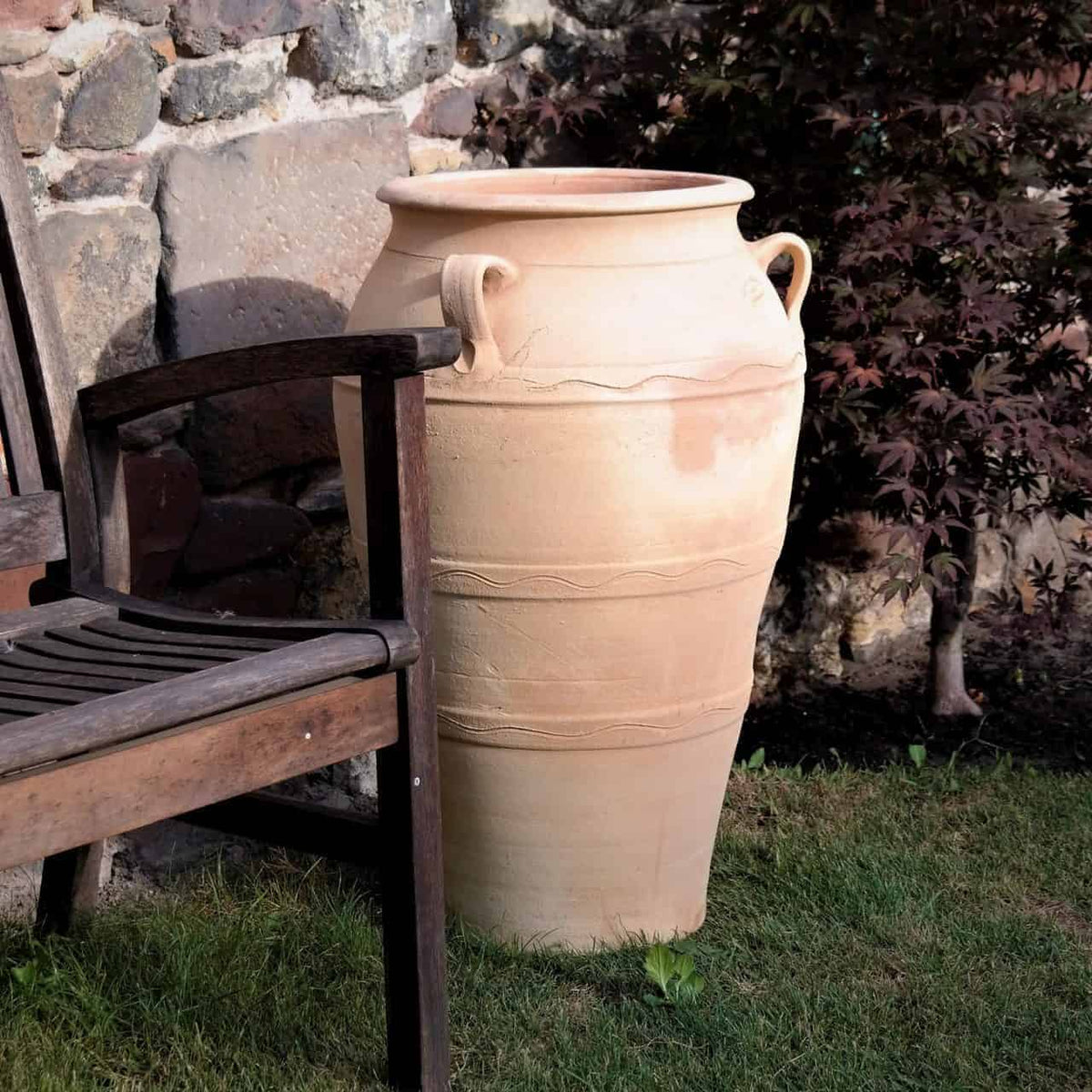 Eine große beige Terrakotta-Amphore für den Garten, „Olea“ H=70 cm, mit dekorativen Griffen steht auf Gras neben einem Holzstuhl. Im Hintergrund sorgen eine Steinmauer und eine Olea-Pflanze mit dunklen Blättern für eine ruhige Gartenszene.