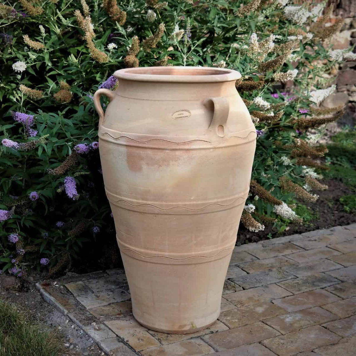 Die Gartenamphore „Olea“, eine große beige Keramikvase mit einer Höhe von 70 cm, zeichnet sich durch horizontale dekorative Linien und eine verwitterte Optik aus. Sie steht auf einer Ziegelfläche zwischen üppigem grünem Laub und violetten Blüten und harmoniert harmonisch mit ihren frostbeständigen Begleitern.