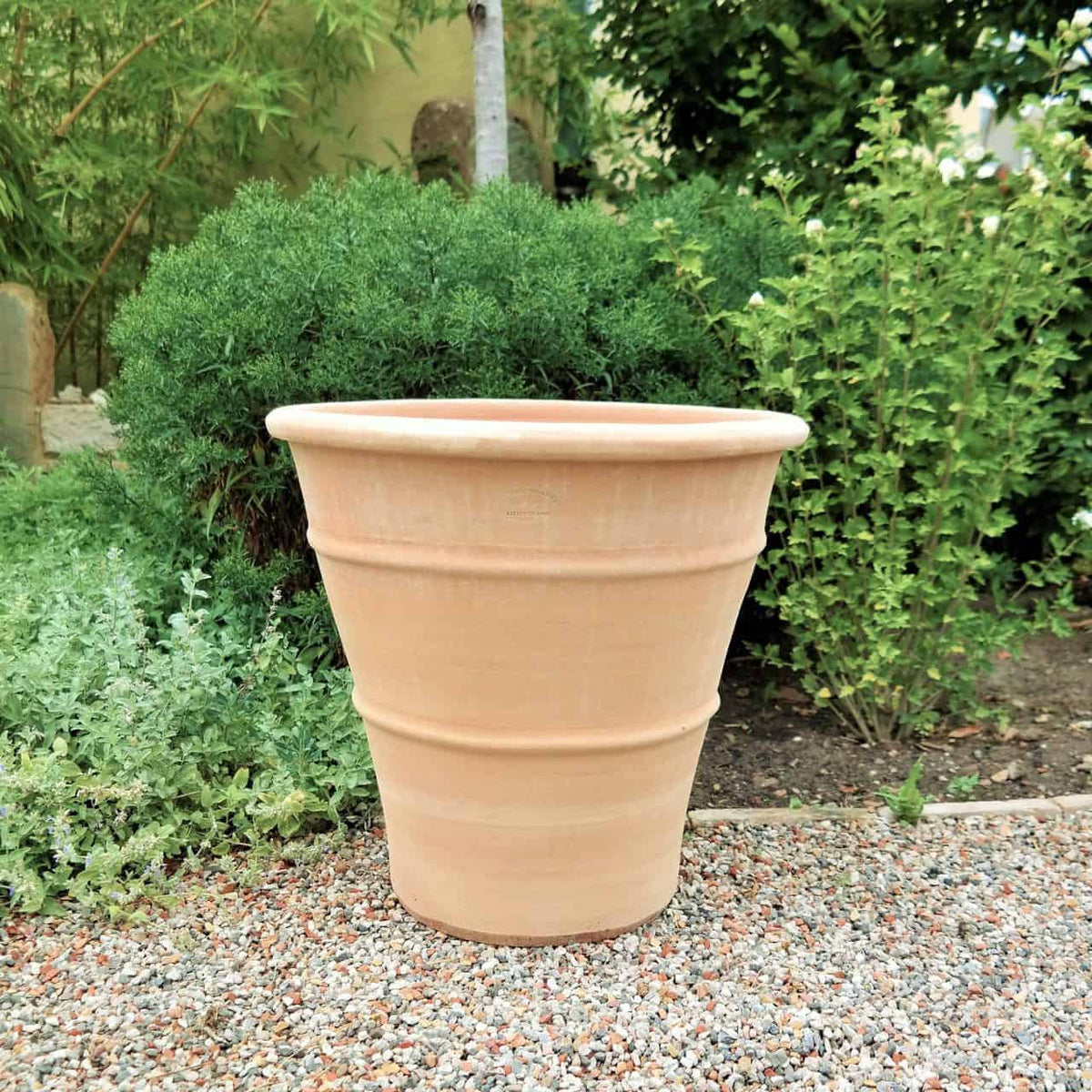 Ein frostfester Terrakotta-Pflanzkübel „Hibiskus“ steht auf einer Kiesfläche in einer Gartenumgebung. Der große, beige Topf mit einer Höhe von 50 cm ist leer und von üppigem grünem Blattwerk und verschiedenen Pflanzen umgeben, darunter leuchtende Hibiskusblüten.