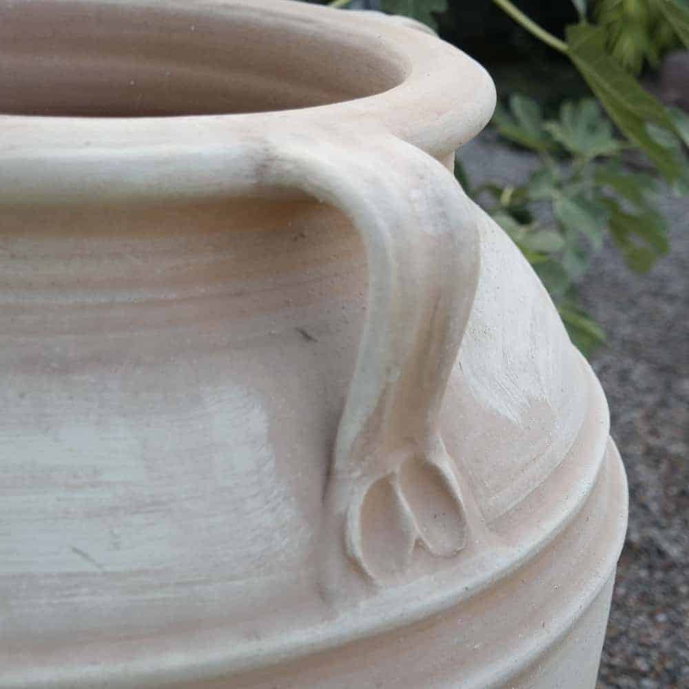 Nahaufnahme der frostfesten Terrakotta-Amphore „Datura“, einem beigen Keramiktopf mit Henkel, 70 cm hoch. Der Topf hat eine glatte, leicht strukturierte Oberfläche mit dezenten horizontalen Rillen. Im unscharfen Hintergrund ist eine grünblättrige Datura-Pflanze zu sehen, und die Szenerie scheint im Freien auf einer Kiesfläche zu stehen.