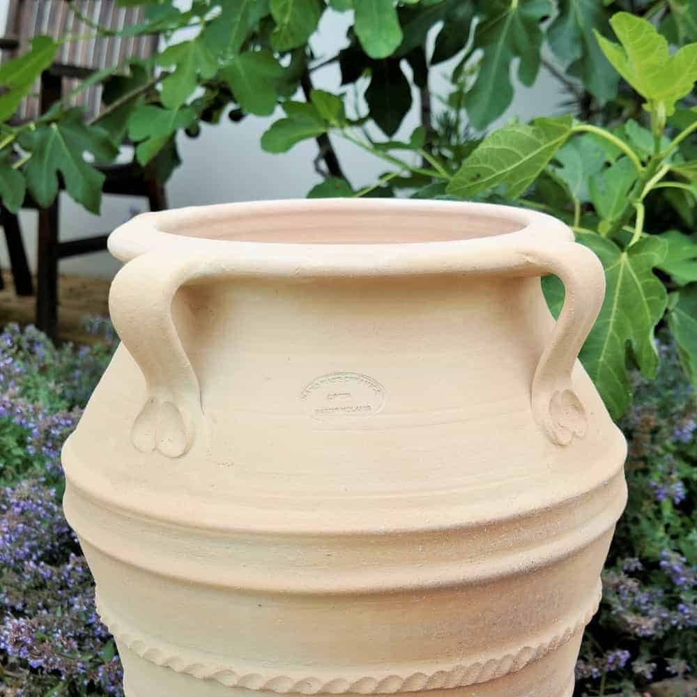Eine große frostfeste Terrakotta-Amphore mit Henkel "Datura", H=50 cm, mit rauer, natürlicher Oberfläche steht in einer Gartenumgebung. Die Amphore ist teilweise von grünem Laub umgeben, im Hintergrund ist ein Stuhl zu sehen.