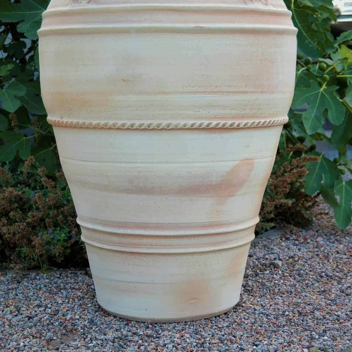 Eine große, erdfarbene, frostfeste Terrakotta-Amphore „Datura“, 90 cm hoch, mit horizontalen Rillen und dezenten seilartigen Verzierungen, steht auf einer Kiesfläche. Im unscharfen Hintergrund sind grüne Datura-Pflanzen mit Blättern zu sehen.