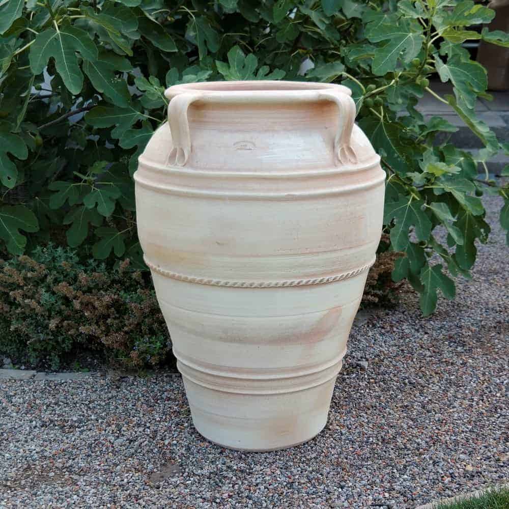 Eine große, beige, frostbeständige Terrakotta-Amphore „Datura“ H= 90 cm mit zwei Griffen steht auf einer Kiesfläche, umgeben von üppigem grünem Blattwerk, darunter blühender Datura. Die Amphore hat ein strukturiertes Band um ihren Mittelteil und verjüngt sich leicht zur Basis hin, wodurch ein traditionelles, rustikales Design entsteht.