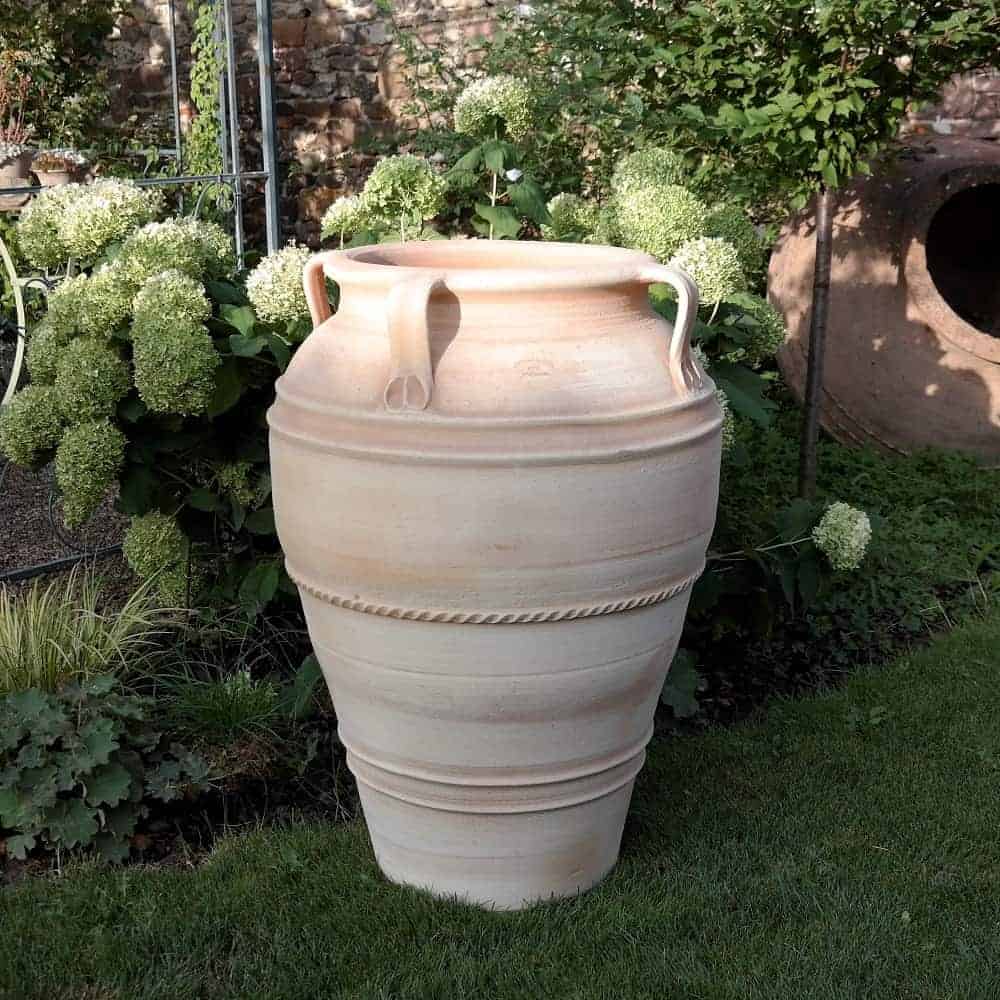 Eine große, beige, handgefertigte Terrakotta-Amphore „Datura“ ist frostsicher und 90 cm hoch und steht im Freien auf einer Rasenfläche. Um den Topf herum stehen üppig blühende Pflanzen mit weißen Blüten und schaffen eine ruhige Gartenszene.