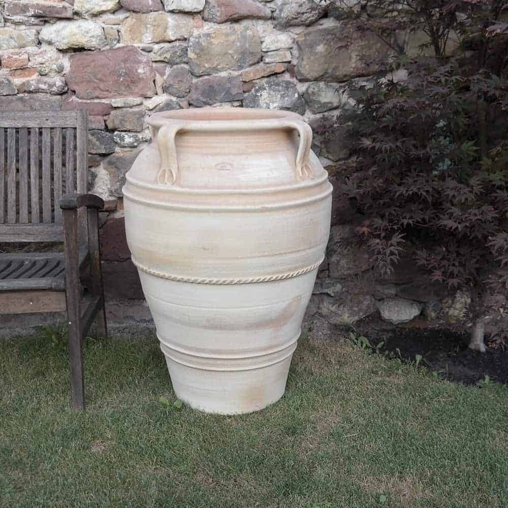 Die große frostfeste Terrakotta-Amphore „Datura“ H=90 cm, eine große beige Amphore mit zwei Henkeln und dekorativen Rillen, steht auf einer Rasenfläche neben einer Holzbank. Eine Steinmauer in verschiedenen Braun- und Beigetönen dient als Hintergrund, rechts ist teilweise eine Blattpflanze zu sehen.