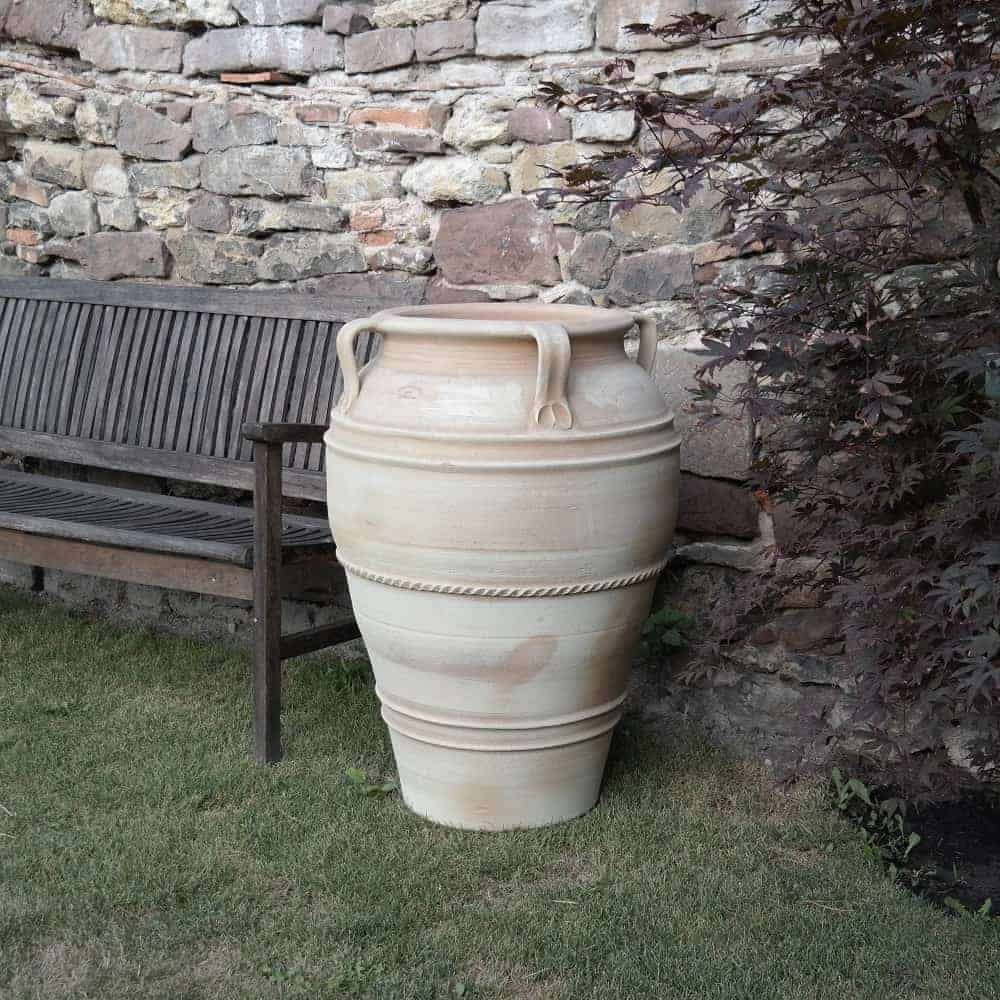 Eine hohe, beige Keramikvase mit dekorativen Griffen und Mustern steht auf dem Rasen neben einer Holzbank. Im Hintergrund ist eine rustikale Steinmauer zu sehen, und auf der rechten Seite ist teilweise eine Blattpflanze zu sehen. In der Nähe ergänzt die große frostfeste Terrakotta-Amphore „Datura“ (Höhe 90 cm) die Szene auf subtile Weise.