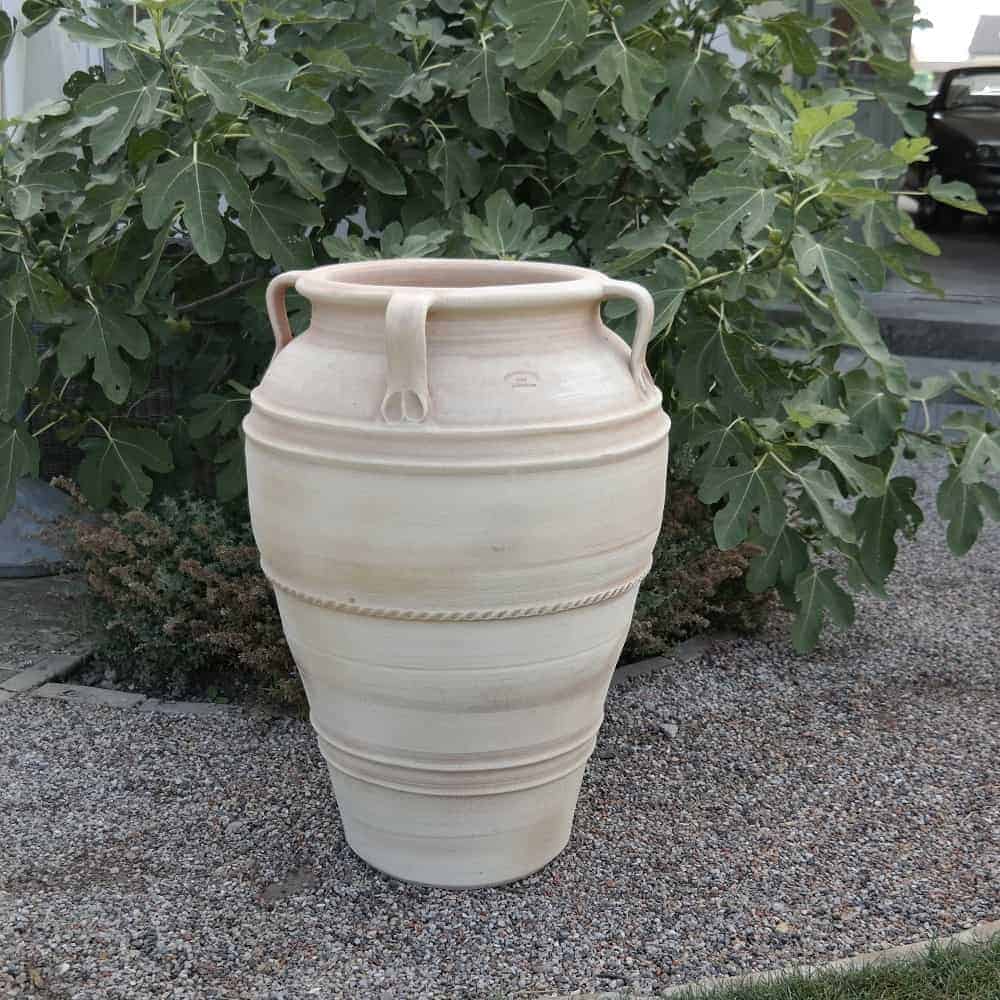 Eine hohe, beige Keramikamphore mit zwei Henkeln steht auf einer Kiesfläche vor üppigem grünem Blattwerk. Die große frostfeste Terrakotta-Amphore „Datura“ H= 90 cm ist mit kreisförmigen Mustern verziert und hat eine schmale Basis, die sich nach oben hin allmählich verbreitert.