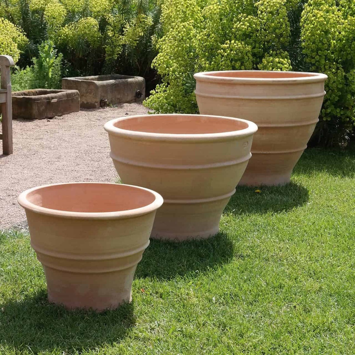 Drei große, leere frostbeständige Terrakotta-Blumentöpfe („Buxus“, 40 cm) unterschiedlicher Größe sind auf einer Rasenfläche vor grünen Buchsbäumen aufgestellt. Die Szene spielt im Freien an einem hellen, sonnigen Tag, im Hintergrund ist ein Kiesweg zu sehen.