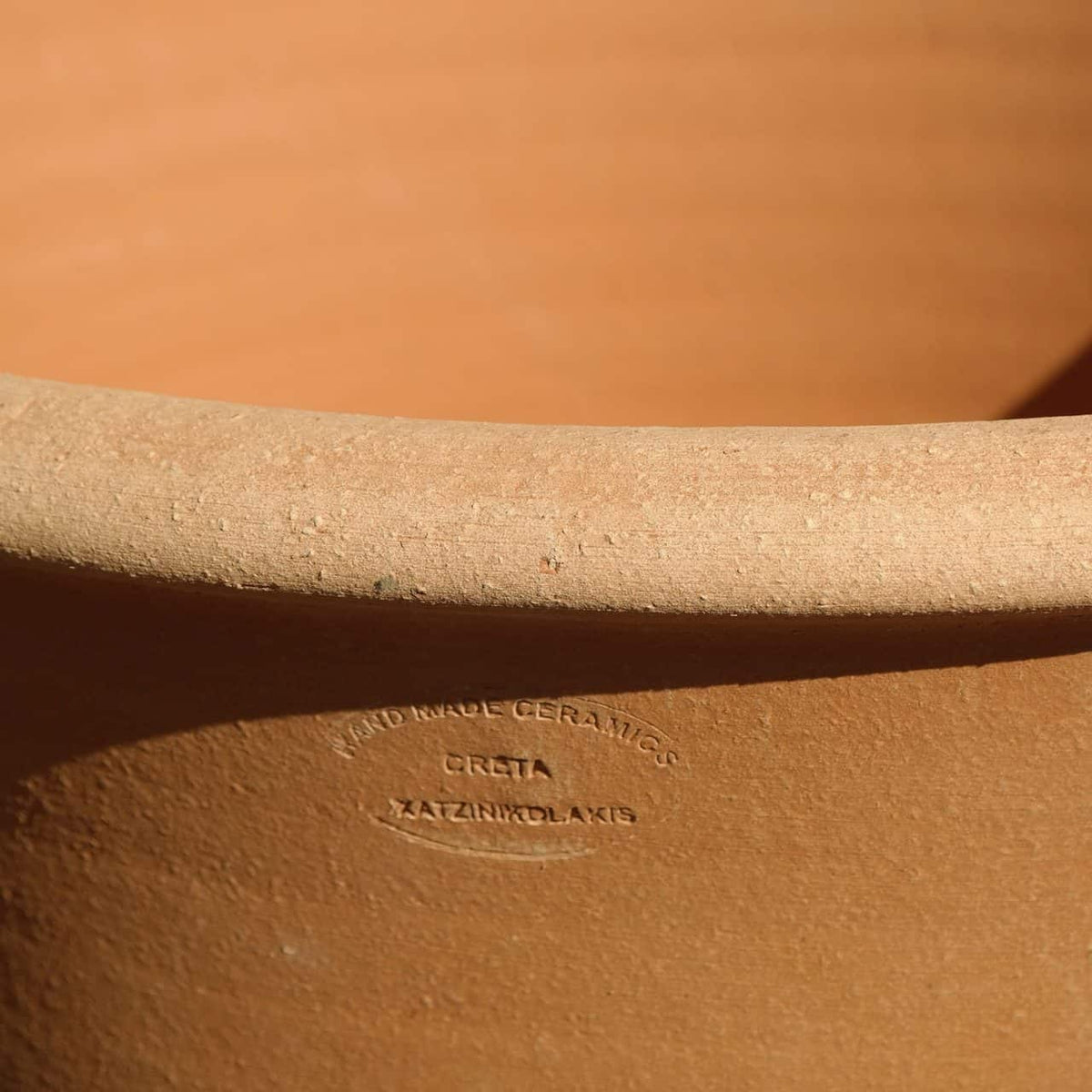 Nahaufnahme eines großen Terrakotta-Keramikgefäßes. Der Rand zeigt einen eingravierten Stempel mit der Aufschrift „HANDGEMACHTE KERAMIK – CRETA – KAZINCLOLAKI“. Die Textur ist rau und unglasiert, was die Handwerkskunst des Stücks unterstreicht.