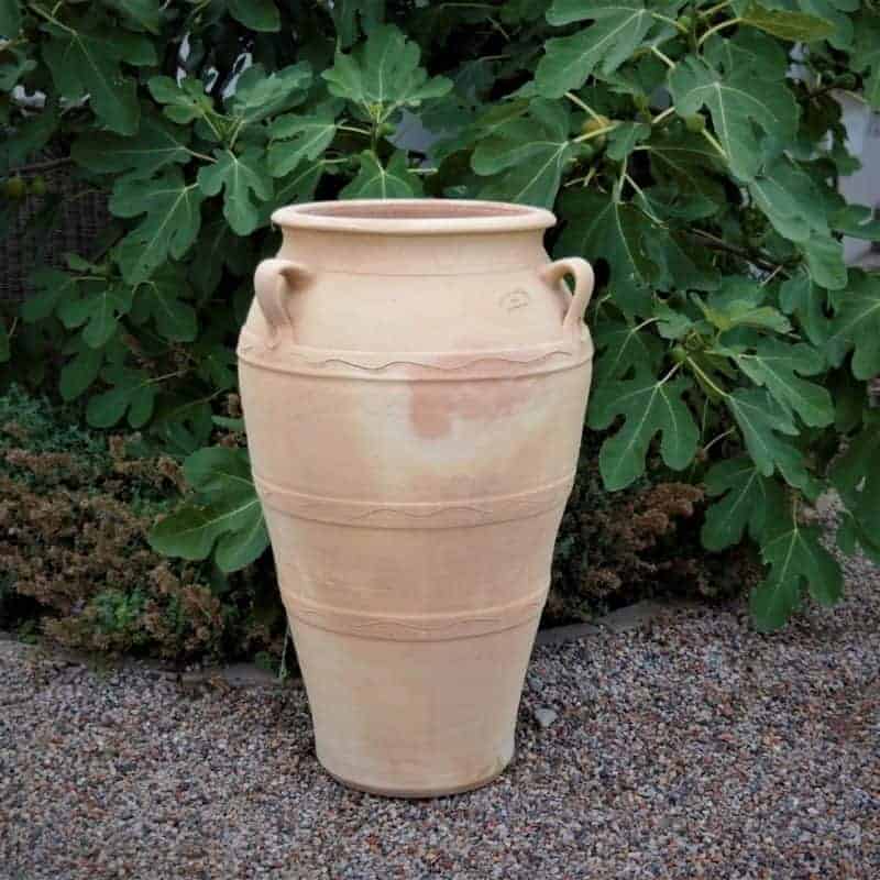 Die Amphore für den Garten "Olea" H=70 cm ist ein hoher, hellbrauner Terrakotta-Topf mit zwei kleinen Griffen, der auf Kies ruht und von üppigem Blattwerk umgeben ist. Dieses frostbeständige Stück hat einen glatten, konischen Körper mit dezenten Bändern und einer weiten Öffnung.