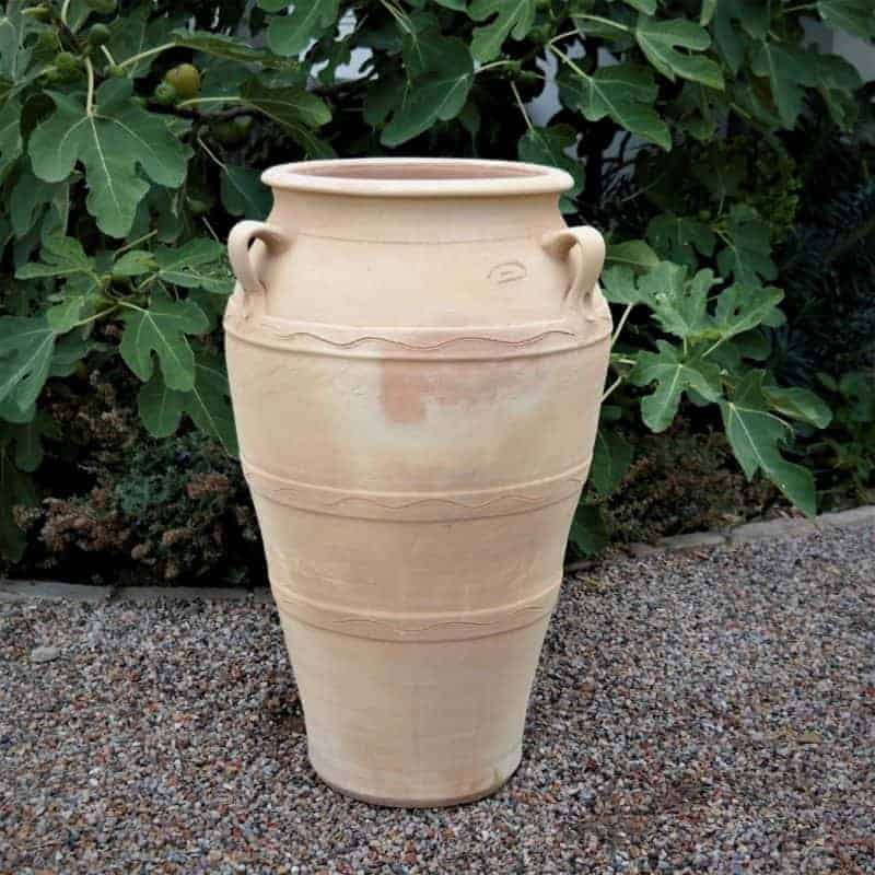 Die 70 cm hohe Amphore für den Garten „Olea“ mit ihrer großen unglasierten Terrakottaform und den dekorativen Linien steht anmutig auf einem Kiesweg. Ihre beiden kleinen seitlichen Henkel ergänzen die üppigen grünen Olea-Blätter im Hintergrund und schaffen eine zauberhafte frostfeste Gartenszene.