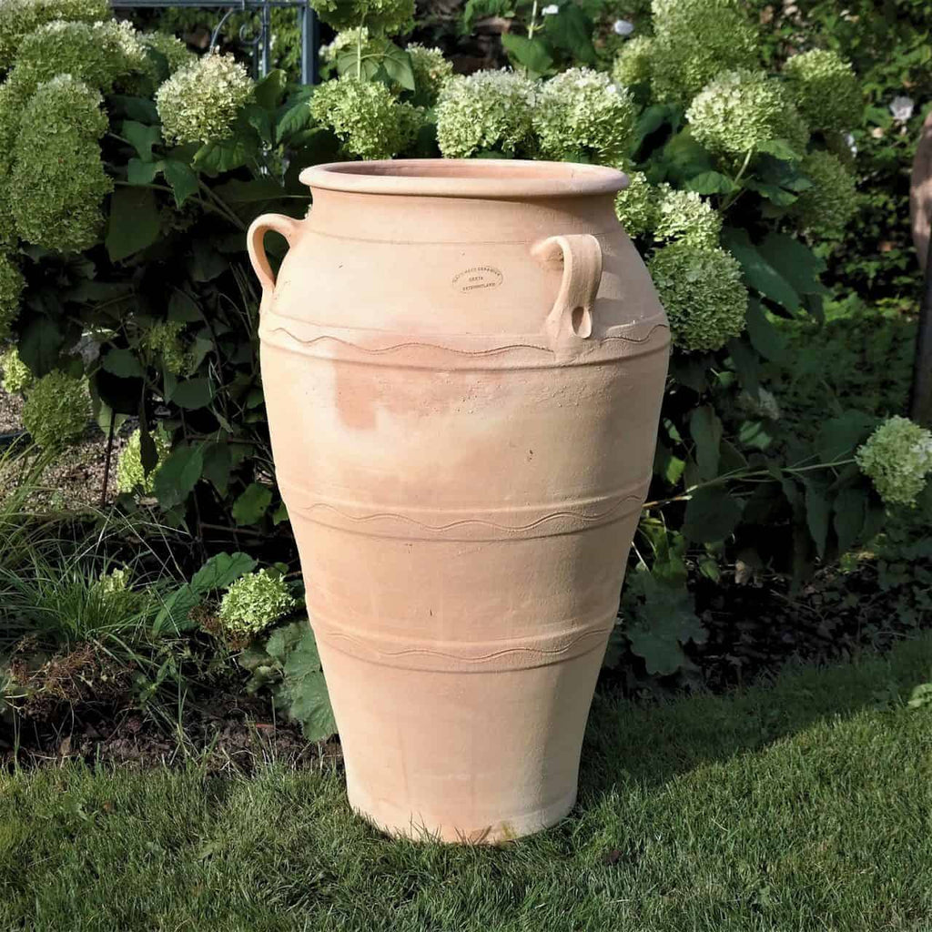 Die hellbeige, 70 cm hohe Gartenamphore „Olea“ steht auf einer Rasenfläche, umgeben von üppigen Hortensien. Die frostbeständige Urne verfügt über zwei Henkel, eine strukturierte Oberfläche mit dezenten horizontalen Linien und eine breite Öffnung.