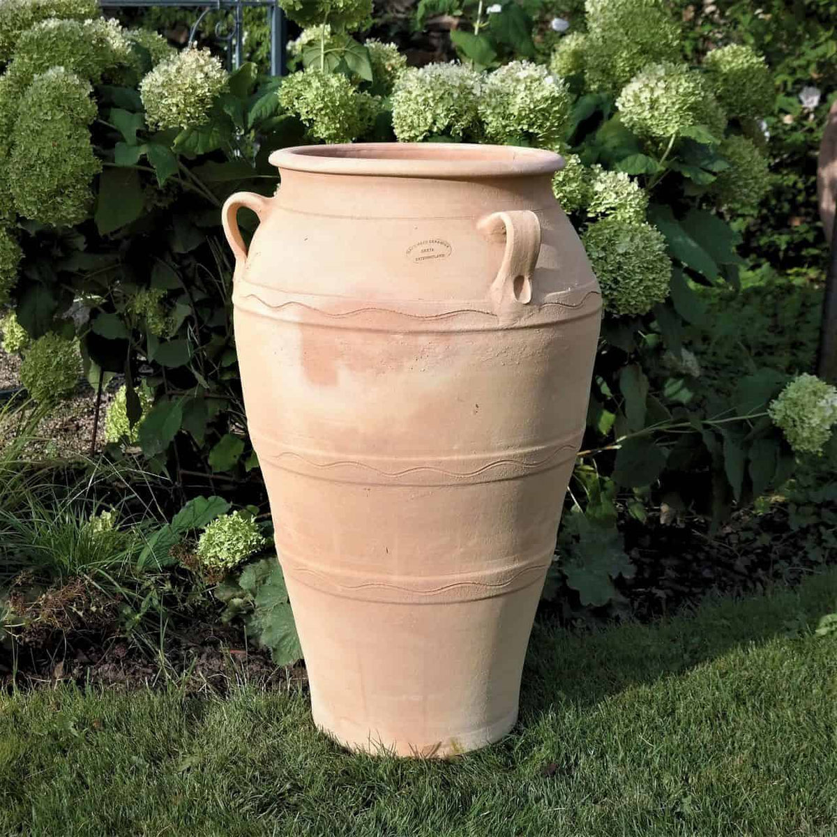 Die hellbeige, 70 cm hohe Gartenamphore „Olea“ steht auf einer Rasenfläche, umgeben von üppigen Hortensien. Die frostbeständige Urne verfügt über zwei Henkel, eine strukturierte Oberfläche mit dezenten horizontalen Linien und eine breite Öffnung.