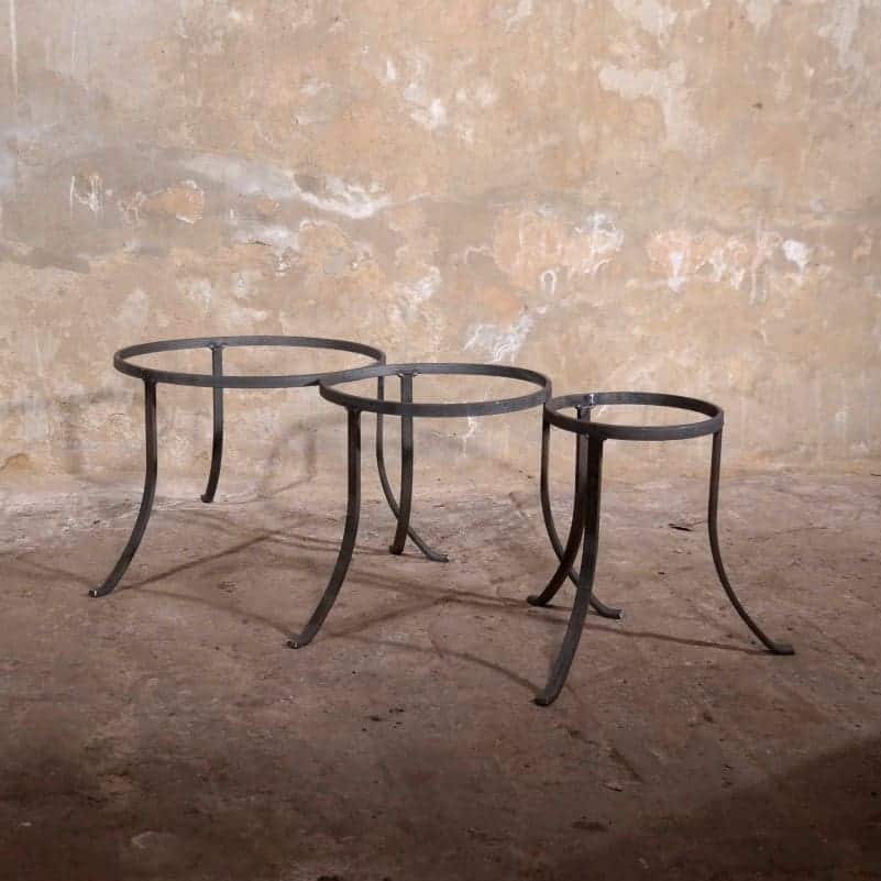 Drei runde, minimalistische Beistelltische aus Metall mit geschwungenen Beinen stehen nebeneinander auf einem Betonboden vor einer strukturierten, abgenutzten Wand. Die Tische haben leere Rahmen ohne Tischplatten.