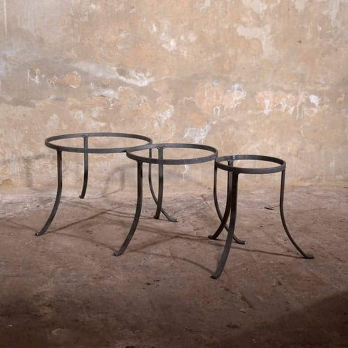 Drei runde, minimalistische Beistelltische aus Metall mit geschwungenen Beinen stehen nebeneinander auf einem Betonboden vor einer strukturierten, abgenutzten Wand. Die Tische haben leere Rahmen ohne Tischplatten.