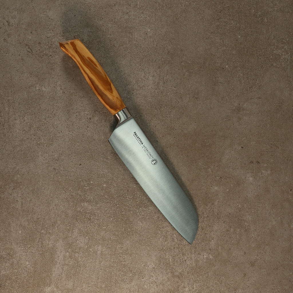 Ein Palatina Werkstatt Messerserie kl.Santokumesser mit einer 16 cm langen Klinge aus rostfreiem Stahl und einem Olivenholzgriff liegt auf einer Betonoberfläche. Die Klinge hat einen Markentext in der Nähe des Griffs. Das Messer hat eine breite, flache Klinge und eine sanft geschwungene Kante, die typischerweise zum Hacken und Schneiden verwendet wird.