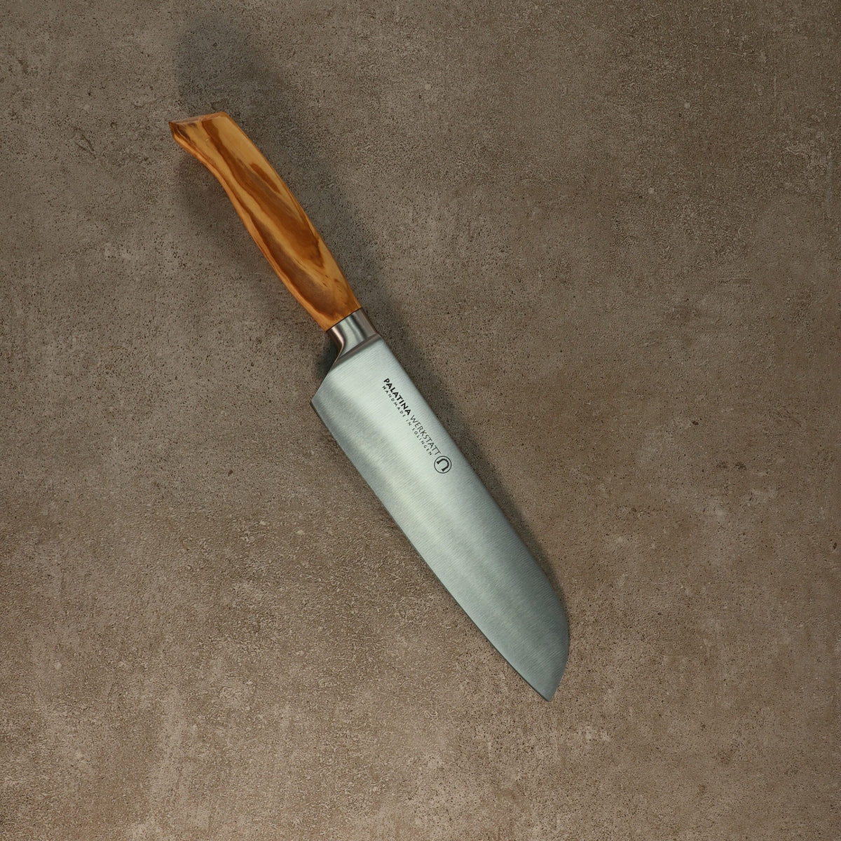 Ein Palatina Werkstatt Messerserie kl.Santokumesser mit einer 16 cm langen Klinge aus rostfreiem Stahl und einem Olivenholzgriff liegt auf einer Betonoberfläche. Die Klinge hat einen Markentext in der Nähe des Griffs. Das Messer hat eine breite, flache Klinge und eine sanft geschwungene Kante, die typischerweise zum Hacken und Schneiden verwendet wird.