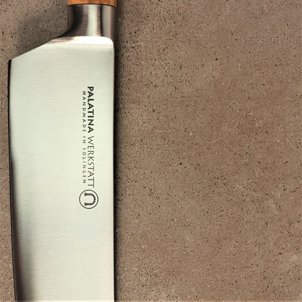 Nahaufnahme eines Küchenmessers mit der Aufschrift „Palatina Werkstatt Handmade in Solingen“ auf der Klinge. Das Palatina Werkstatt Messerserie kl.Santoku 16 cm Klinge rostfrei verfügt über einen eleganten Griff aus Olivenholz und ist mit einer strukturierten, beigen Oberfläche versehen.