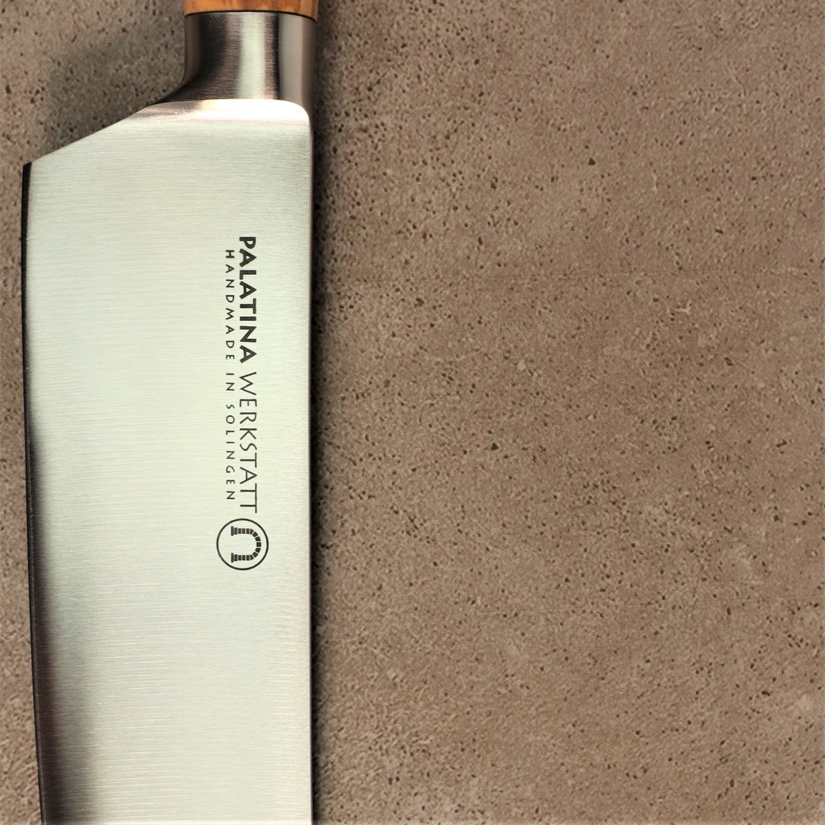 Nahaufnahme eines Küchenmessers mit der Aufschrift „Palatina Werkstatt Handmade in Solingen“ auf der Klinge. Das Palatina Werkstatt Messerserie kl.Santoku 16 cm Klinge rostfrei verfügt über einen eleganten Griff aus Olivenholz und ist mit einer strukturierten, beigen Oberfläche versehen.