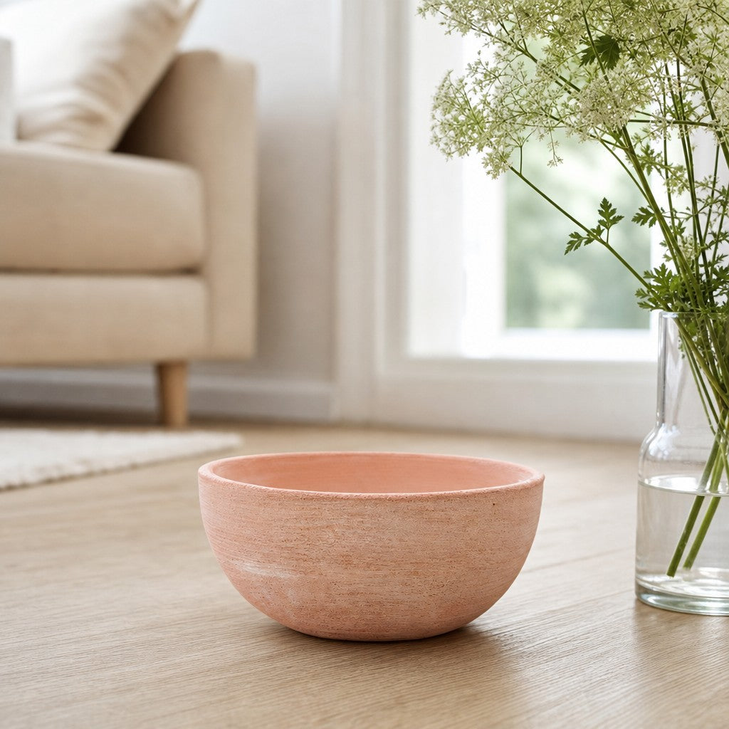 Eine moderne Pflanzschale mit einem Durchmesser von 29–50 cm steht auf einem Holzboden neben einer klaren Vase mit weißen Blumen; im Hintergrund sind ein Sofa und ein großes Fenster bei sanfter Beleuchtung zu sehen.