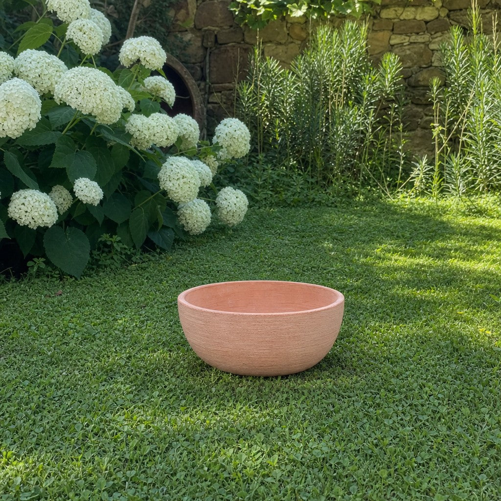 Eine moderne Pflanzschale mit einem Durchmesser von 29–50 cm in Rosa steht auf grünem Gras im Garten, umgeben von weißen Hortensien und Blattpflanzen vor einer Steinmauer.