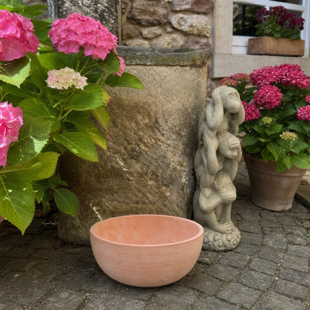 Eine moderne Pflanzschale mit einem Durchmesser von 29–50 cm steht auf dem Kopfsteinpflaster neben einem hohen Steinbrunnen, gepflanzten rosa Hortensien und einer Steinfigur, im Hintergrund sind eine Steinmauer und ein Fenster zu sehen.