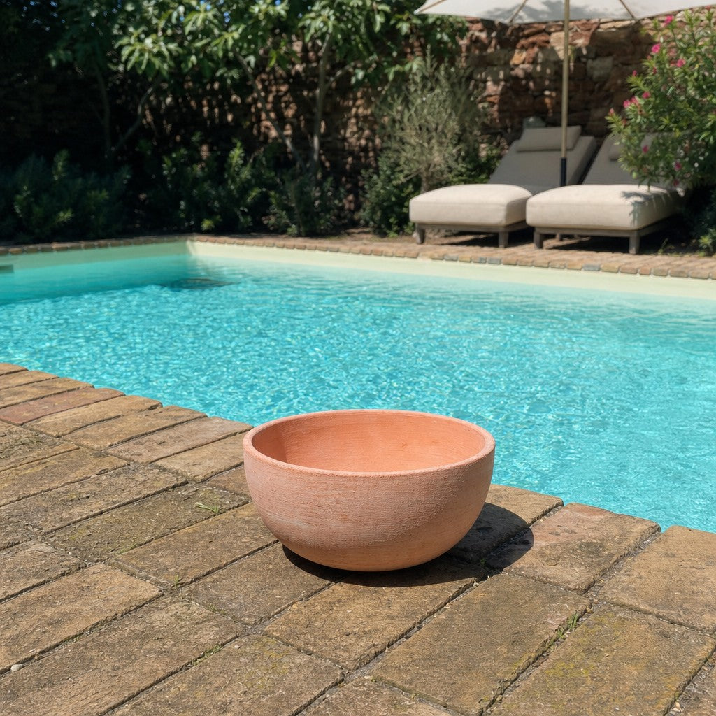 Eine moderne Pflanzschale mit einem Durchmesser von 29–50 cm steht auf einer sonnigen Ziegelterrasse neben einem klaren blauen Pool; im Hintergrund sind zwei gepolsterte Liegestühle und Grünpflanzen zu sehen.