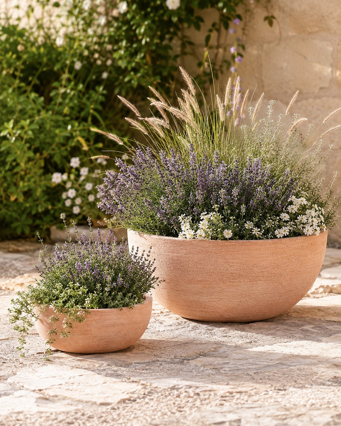 Zwei moderne Pflanzschalen (Durchmesser 29–50 cm) stehen auf einer Steinterrasse und sind mit blühendem Lavendel, weißen Blumen und Ziergräsern bepflanzt. Im Hintergrund sind eine Steinmauer und grünes Laub zu sehen.
