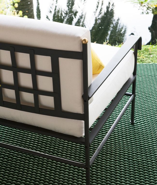 Nahaufnahme eines modernen Outdoor-Sessels Toscana aus Eisen in Graphitgrau mit weißen Kissen, platziert auf einem grünen strukturierten Teppich. Der Sessel steht neben einem Gewässer, im Hintergrund sind Bäume und Gras zu sehen, was die ruhige Atmosphäre der Toskana heraufbeschwört. Auf dem Sessel ist teilweise ein gelbes Kissen zu sehen.